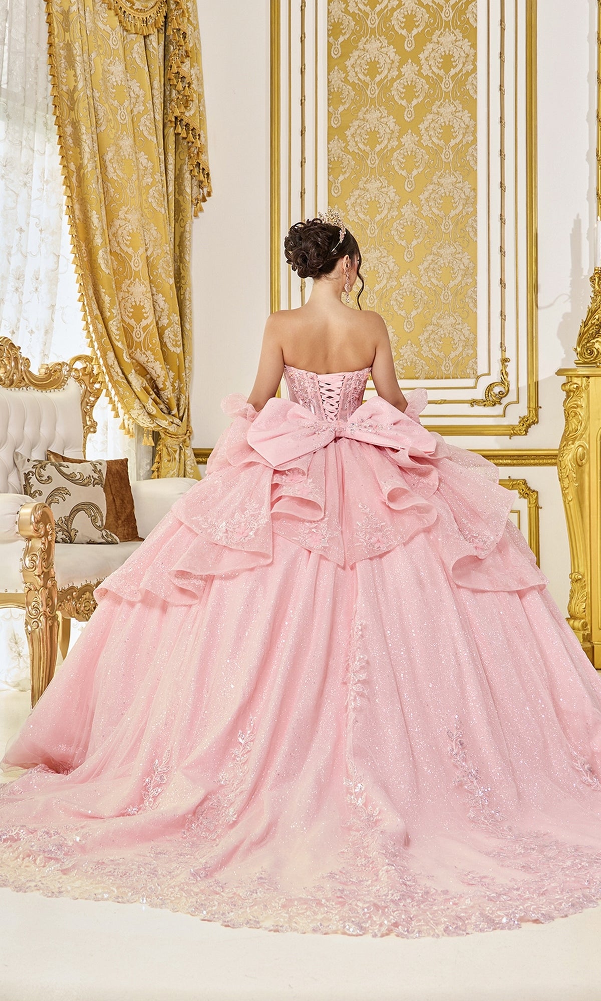 A woman wearing the Quinceanera Dress 5152 by Dancing Queenโa pastel pink, sparkling ball gown with a large bowโstands facing ornate gold and white decor in an elegant room.